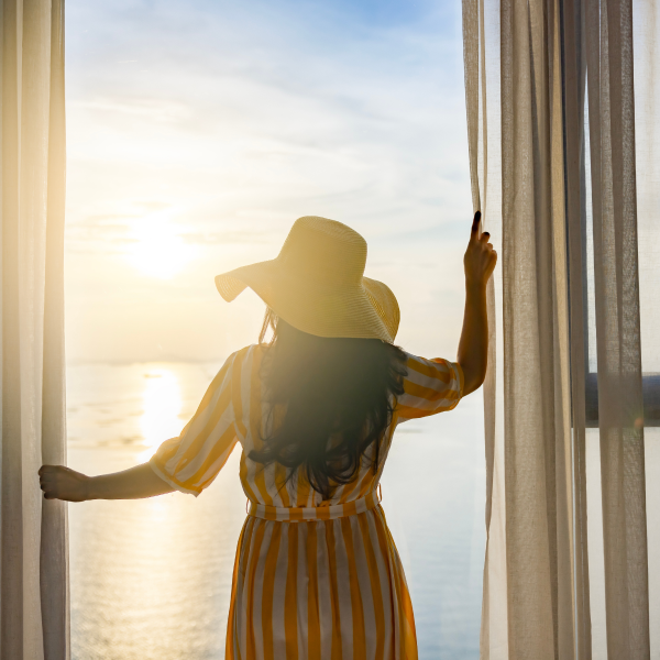 Mujer en vestido amarillo y sombrero mirando por la ventana hacia el océano